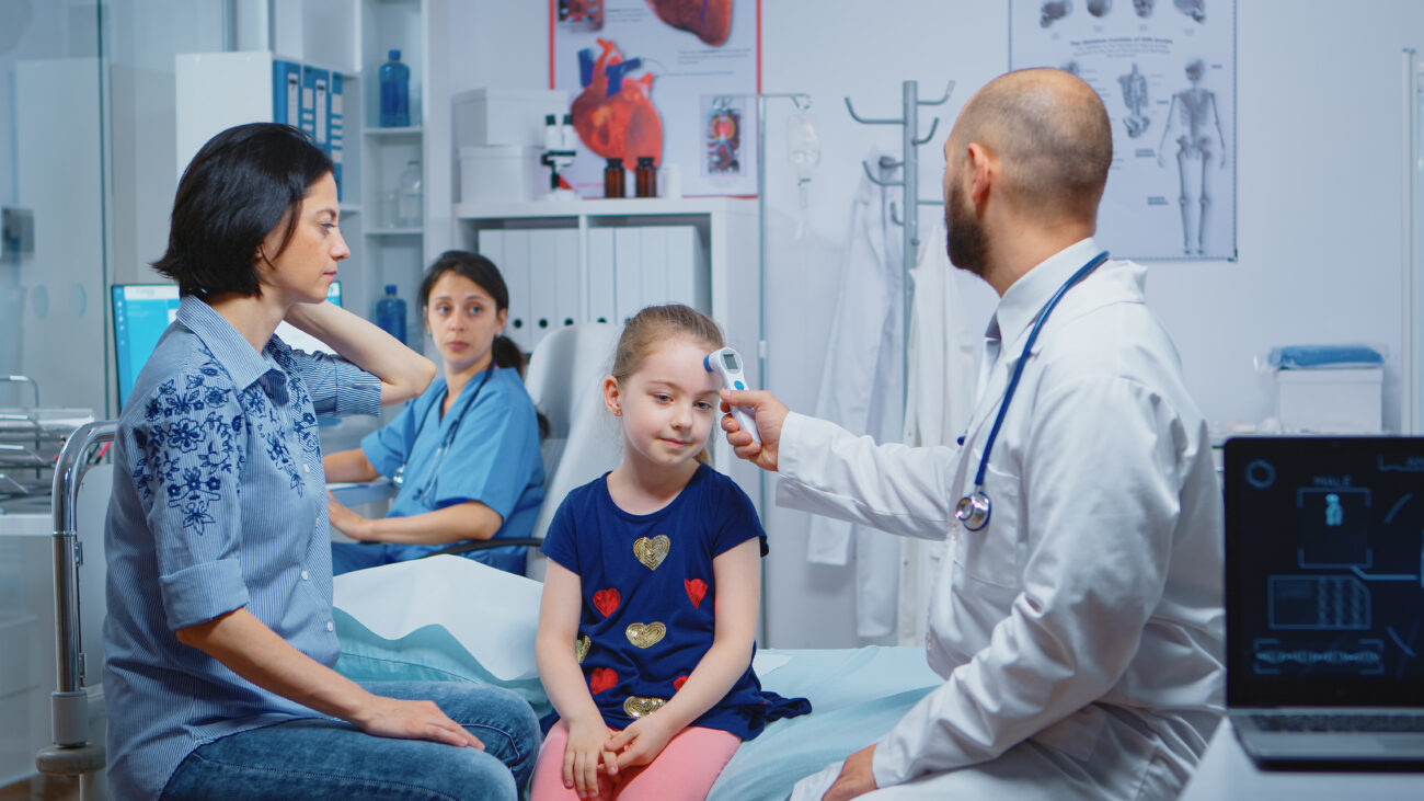 Doctor using a digital thermometer to check the temperature of a young girl in a pediatric hospital room, with her mother and a nurse present.